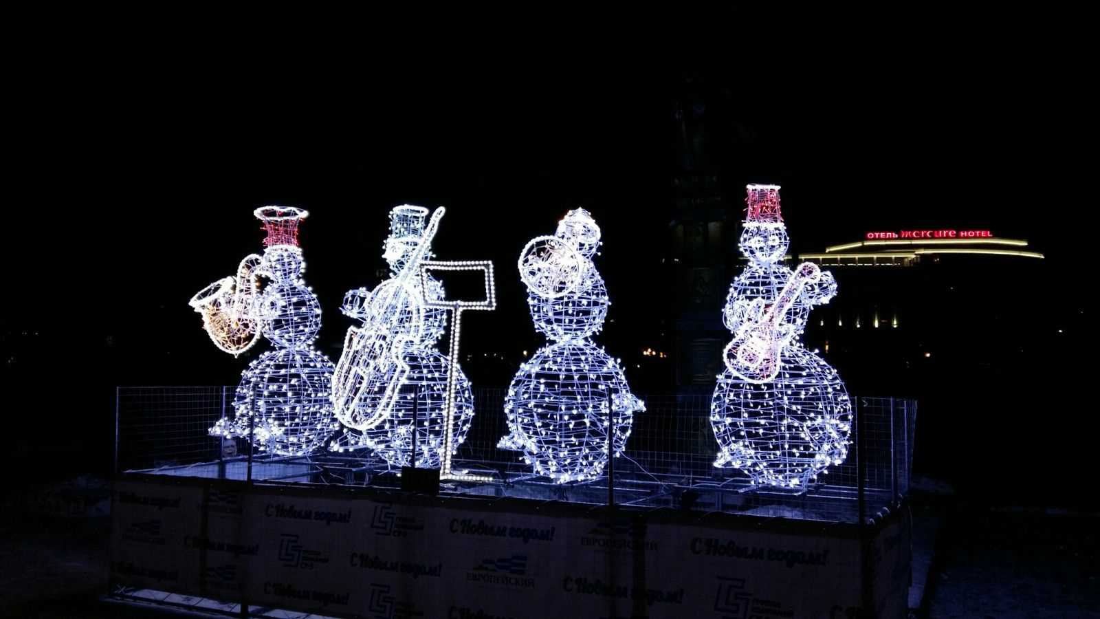 Фотография.  На темном фоне,  в центре стоят 4 фигуры снеговиков – музыкантов. Снеговики сделаны из гирлянд и светятся, у каждого из них свой инструмент . Слева направо – саксофон,  виолончель, труба и гитара. Перед фигурами стоит пюпитр. Снеговики стоят на  невысоком постаменте и огорожены сеткой.  На заднем плане светится вывеска гостиницы «Mercure».